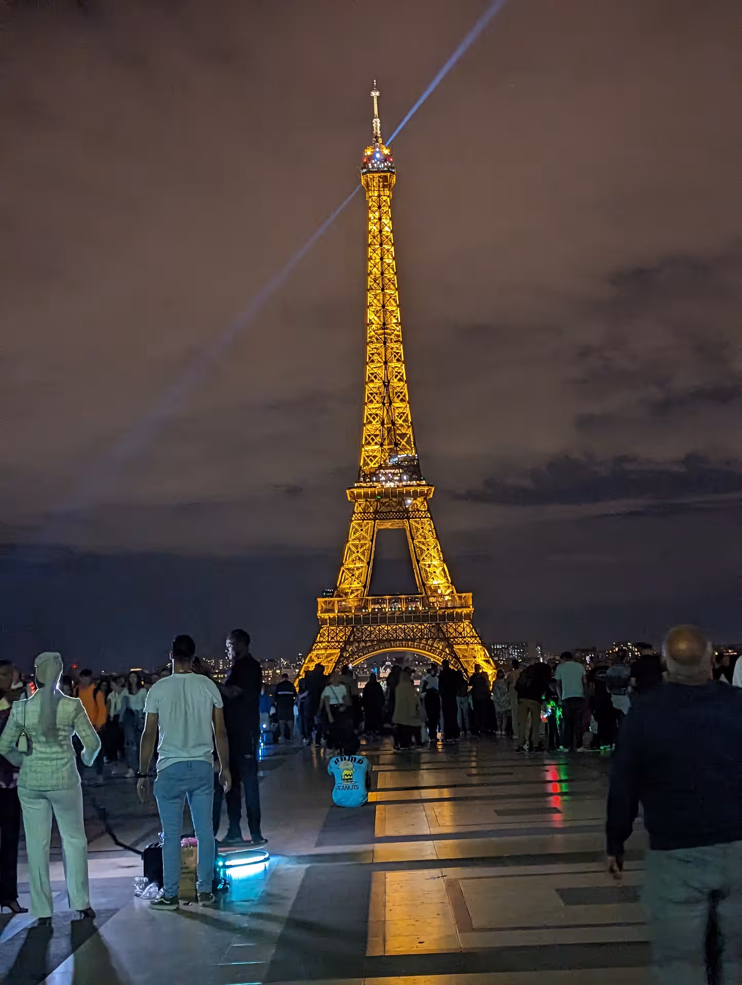 Eiffel Tower illuminated at night with golden lights and summit beacon, viewed from Trocadéro plaza with crowds