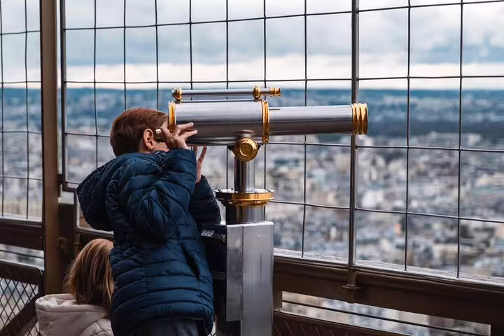 Visitor using coin-operated telescope on Eiffel Tower top floor observation deck overlooking Paris cityscape