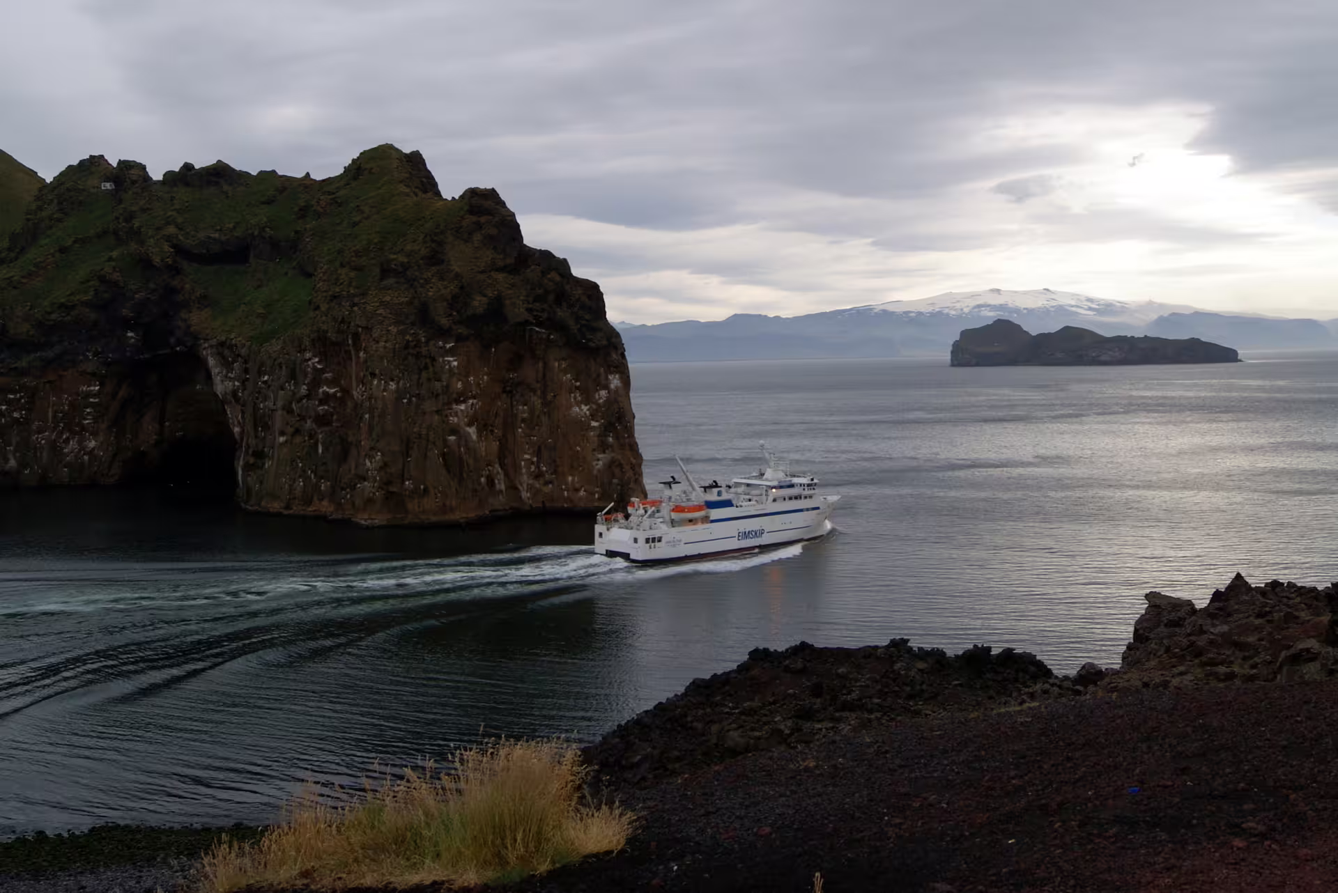 Eimskip ferry sailing past dramatic sea cliffs with snow-capped mountains in South Iceland