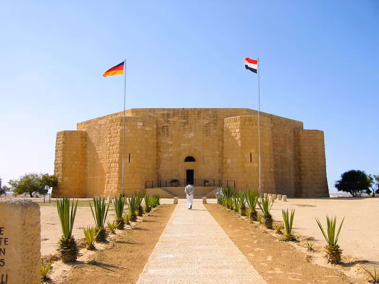 German and Egyptian flags at El Alamein WWII memorial building with stone walls and desert pathway