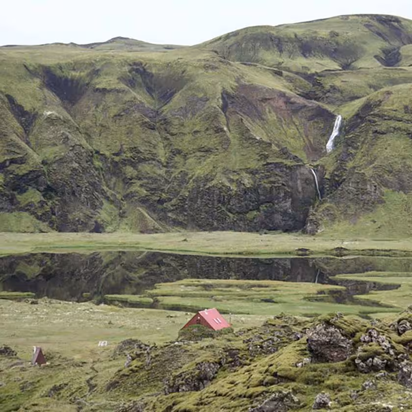 Red mountain hut beside a canyon lake with a waterfall cascading down green mossy cliffs in Iceland