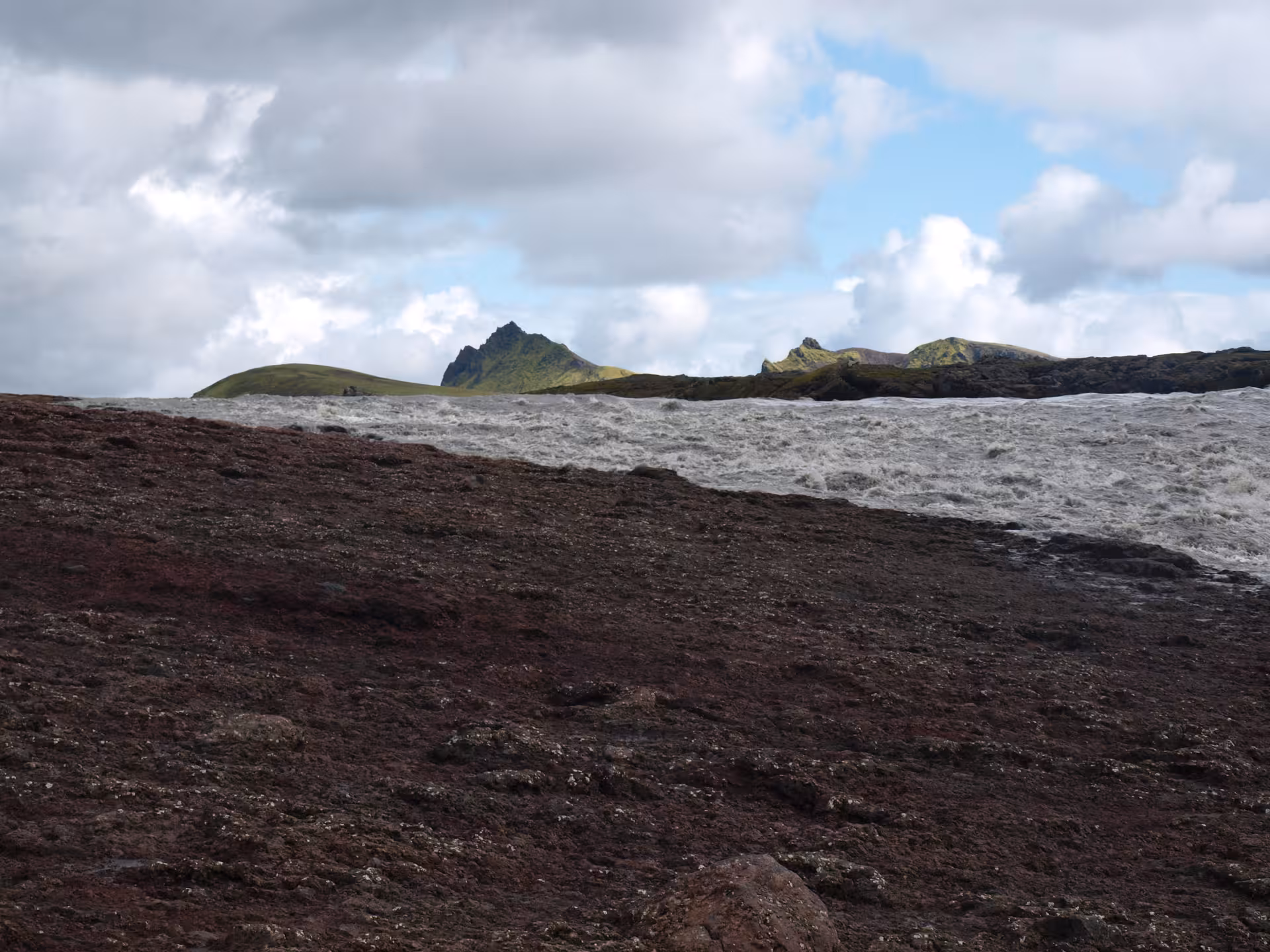 Turbulent glacial river rushing past dark volcanic rocks with green moss-covered peaks in Iceland Highlands