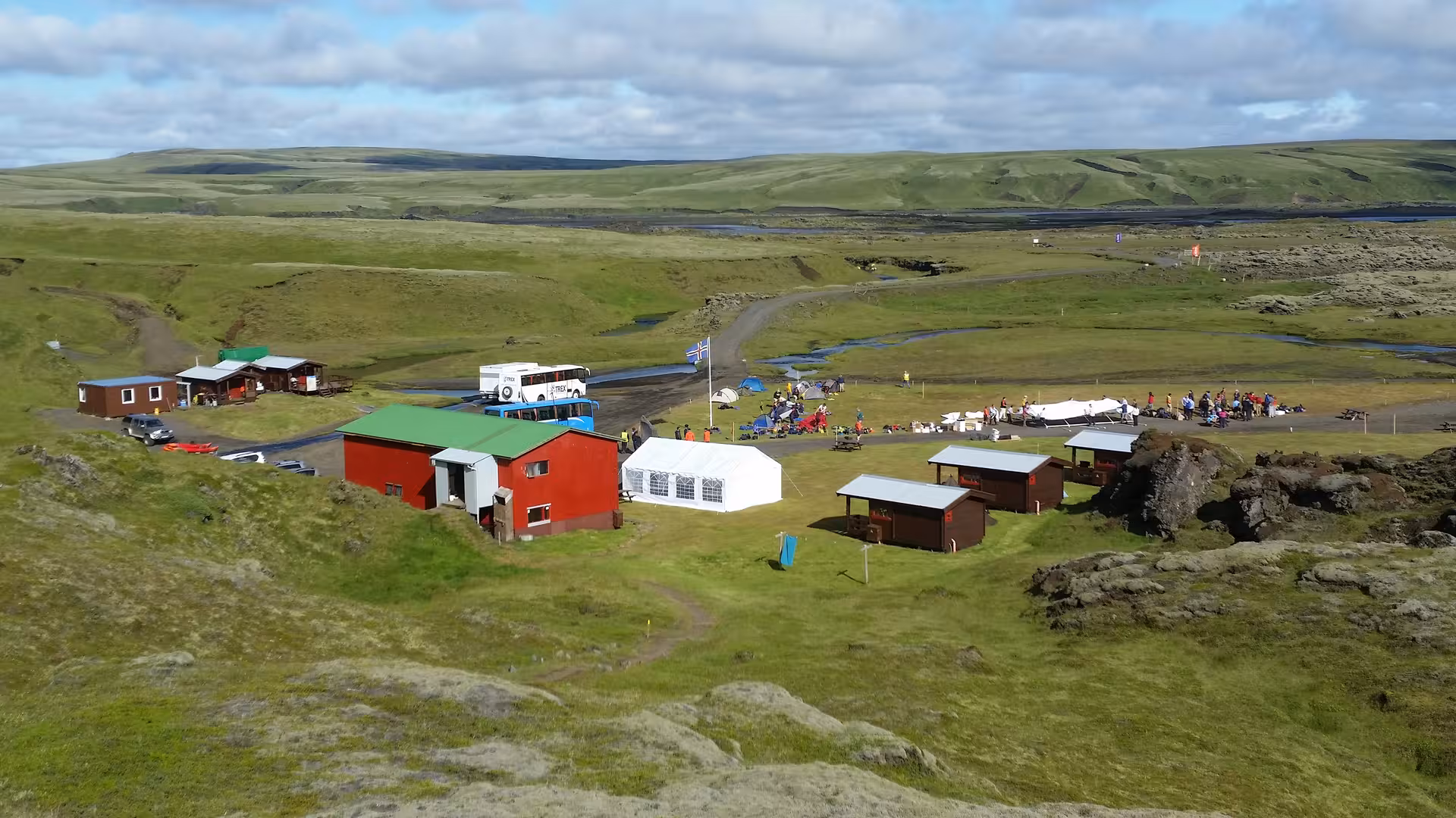 Aerial view of Eldgja highland base camp with red buildings, white tent, and Icelandic flag on green landscape