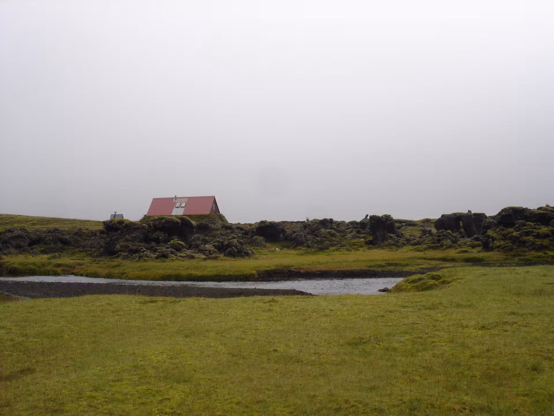 Red-roofed cabin beside a stream in mossy Iceland Highlands lava field on a misty day