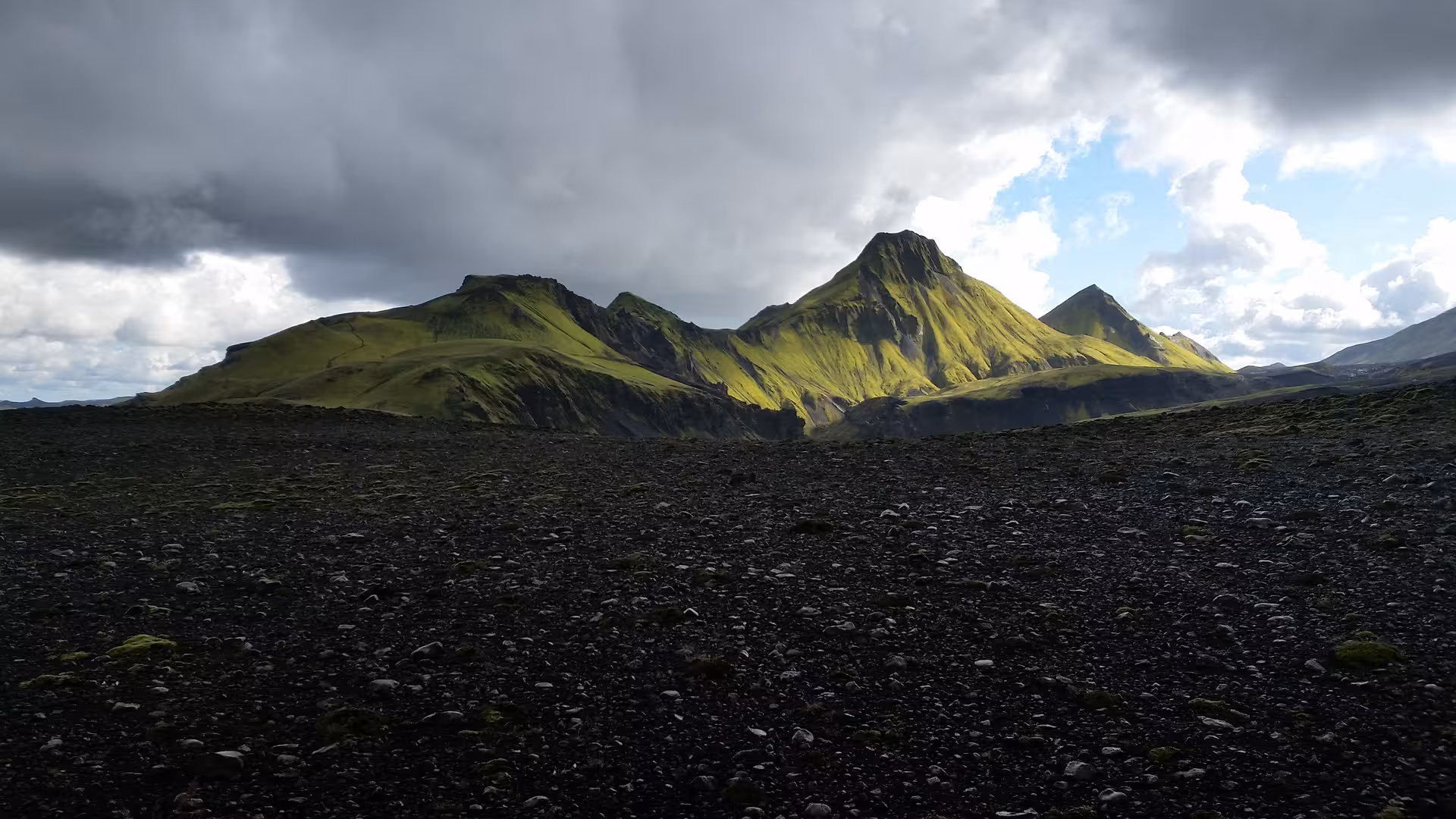 Black volcanic gravel plain with vivid green moss-covered mountains under dramatic storm clouds in Iceland Highlands