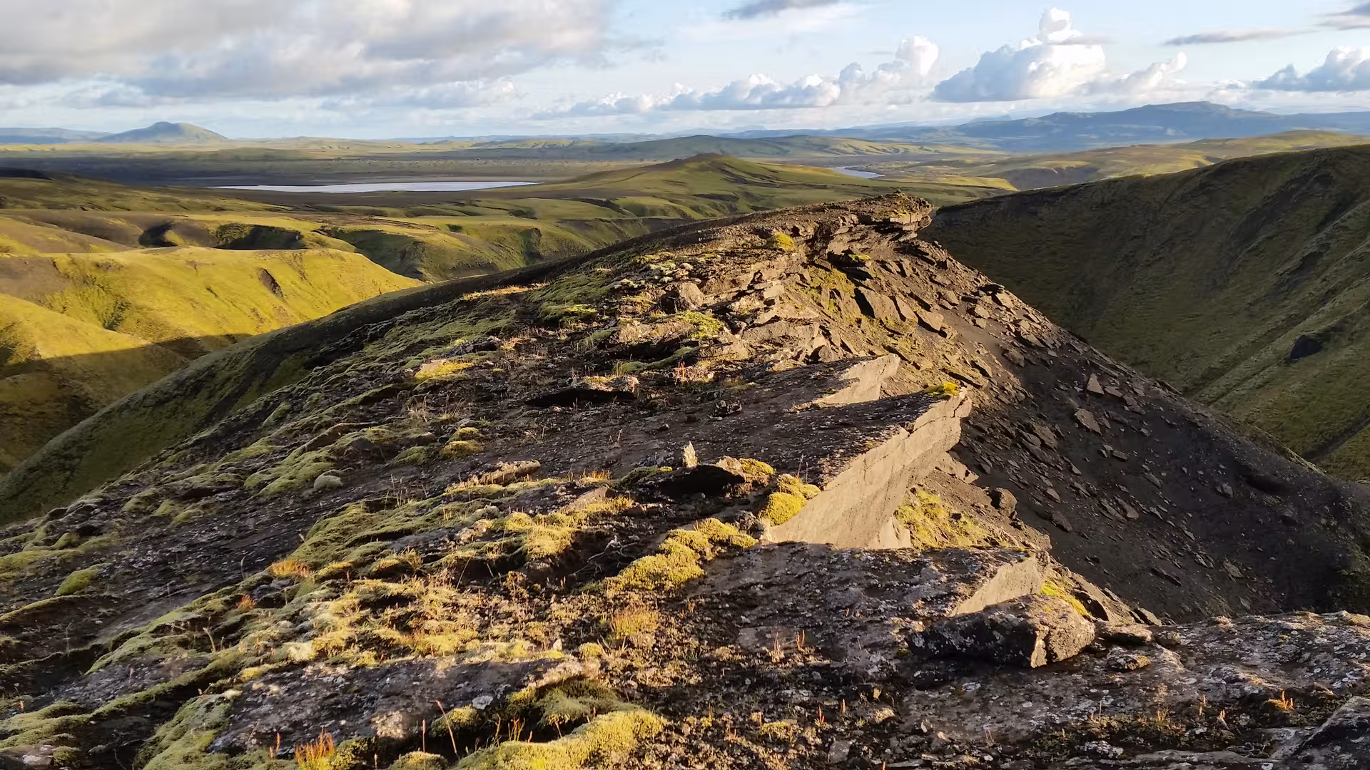 Rocky volcanic ridge trail along Eldgja Canyon with green moss-covered highlands and lakes in background, Iceland