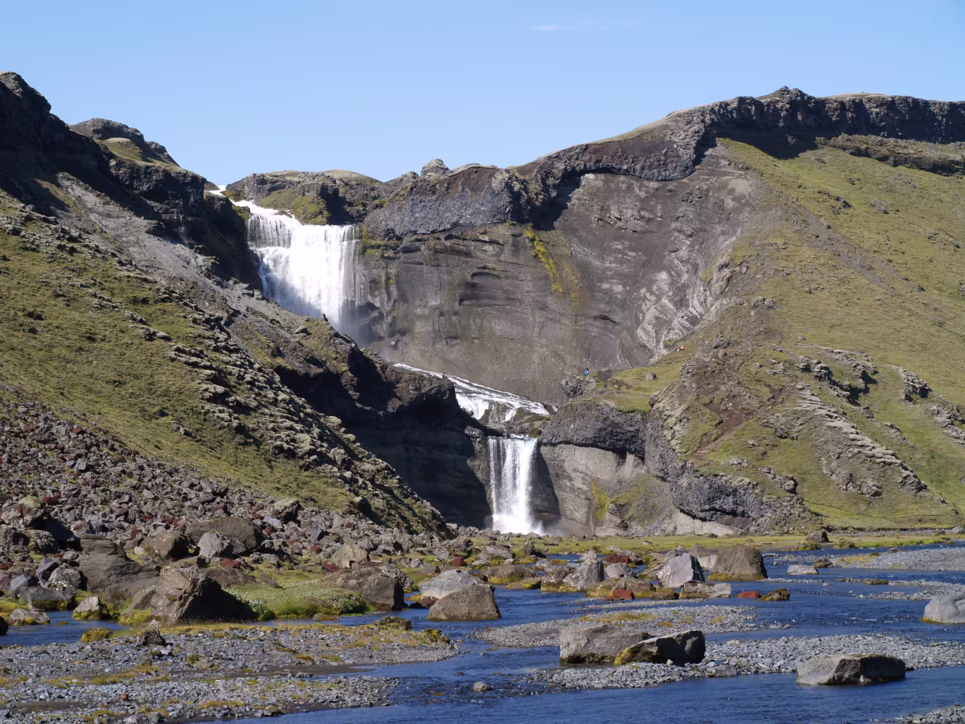 Tiered waterfall cascading down volcanic cliffs into a rocky river in Iceland Highlands
