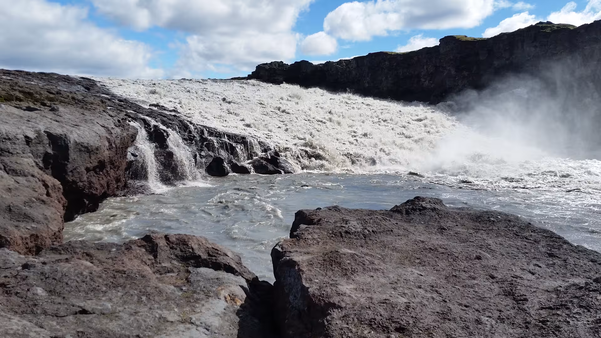 Powerful cascading waterfall crashing over dark volcanic rocks with mist rising under a blue sky in Iceland