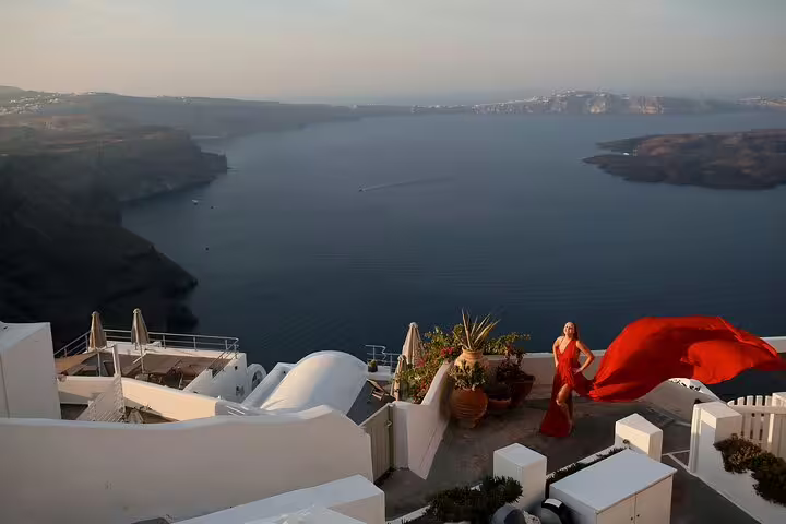Elegant woman in a flowing red dress poses with stunning Santorini caldera views during private photography session.