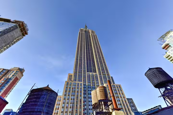 Upward view of Empire State Building towering over NYC rooftops with water towers under blue sky