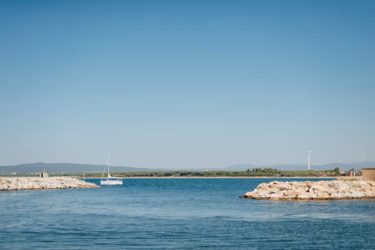 White luxury sailboat gliding through calm Tuscan coastal harbor with rocky breakwaters and clear blue sky.