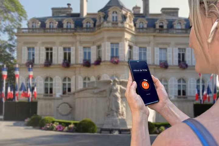 Tourist holding smartphone with GPS audio guide app in front of Épernay Hôtel de Ville with French flags