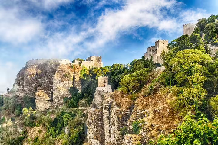 Scenic view of Erice's medieval castles perched on rocky cliffs, surrounded by lush greenery under a vibrant blue sky.
