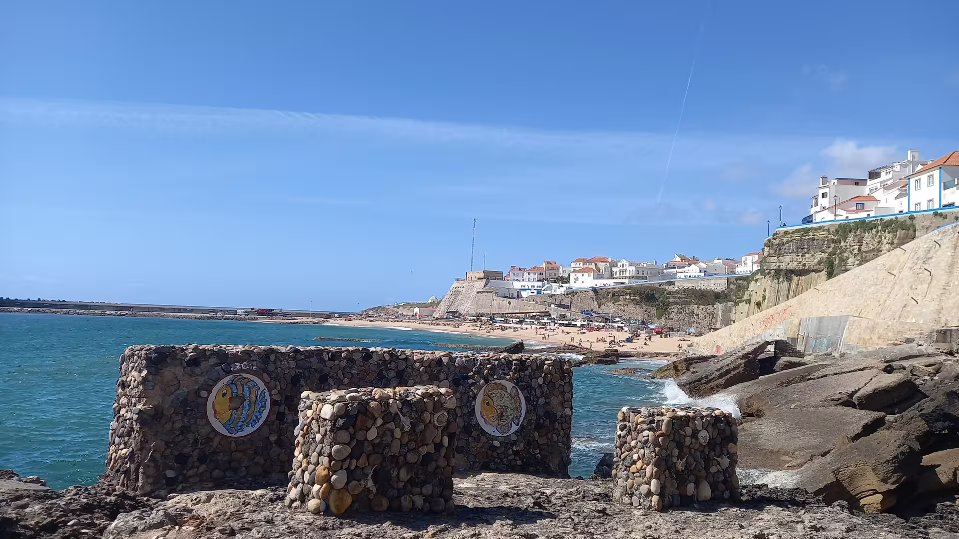 Coastal view of Ericeira beach with scenic stone art and whitewashed buildings under a clear blue sky.