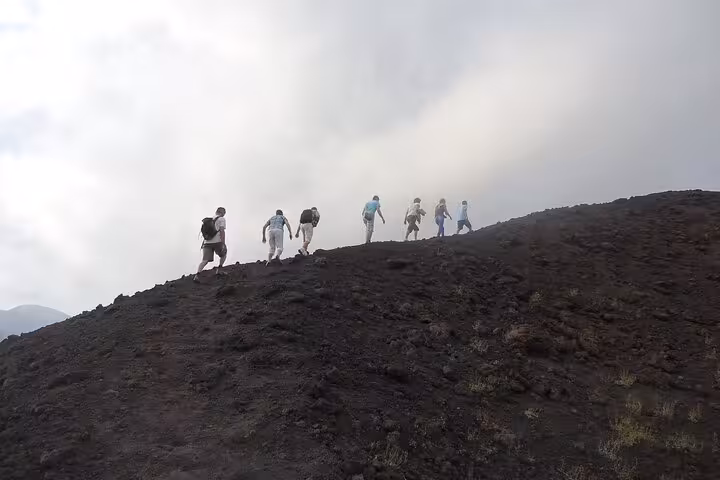 Group of hikers trekking on Mount Etna's rugged volcanic terrain under a cloudy sky, showcasing adventure and exploration.