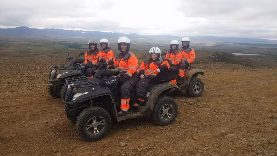 Group of six riders in orange suits and white helmets on ATVs atop a scenic Icelandic wilderness hilltop