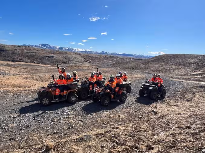Group of riders in orange safety gear on ATV quad bikes on a gravel trail with snowy mountains in background