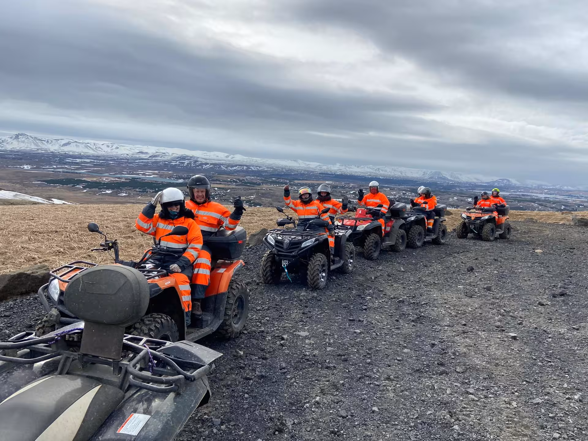 Group of riders in orange safety suits on ATVs lined up on rocky Icelandic highland trail with snowy mountains behind