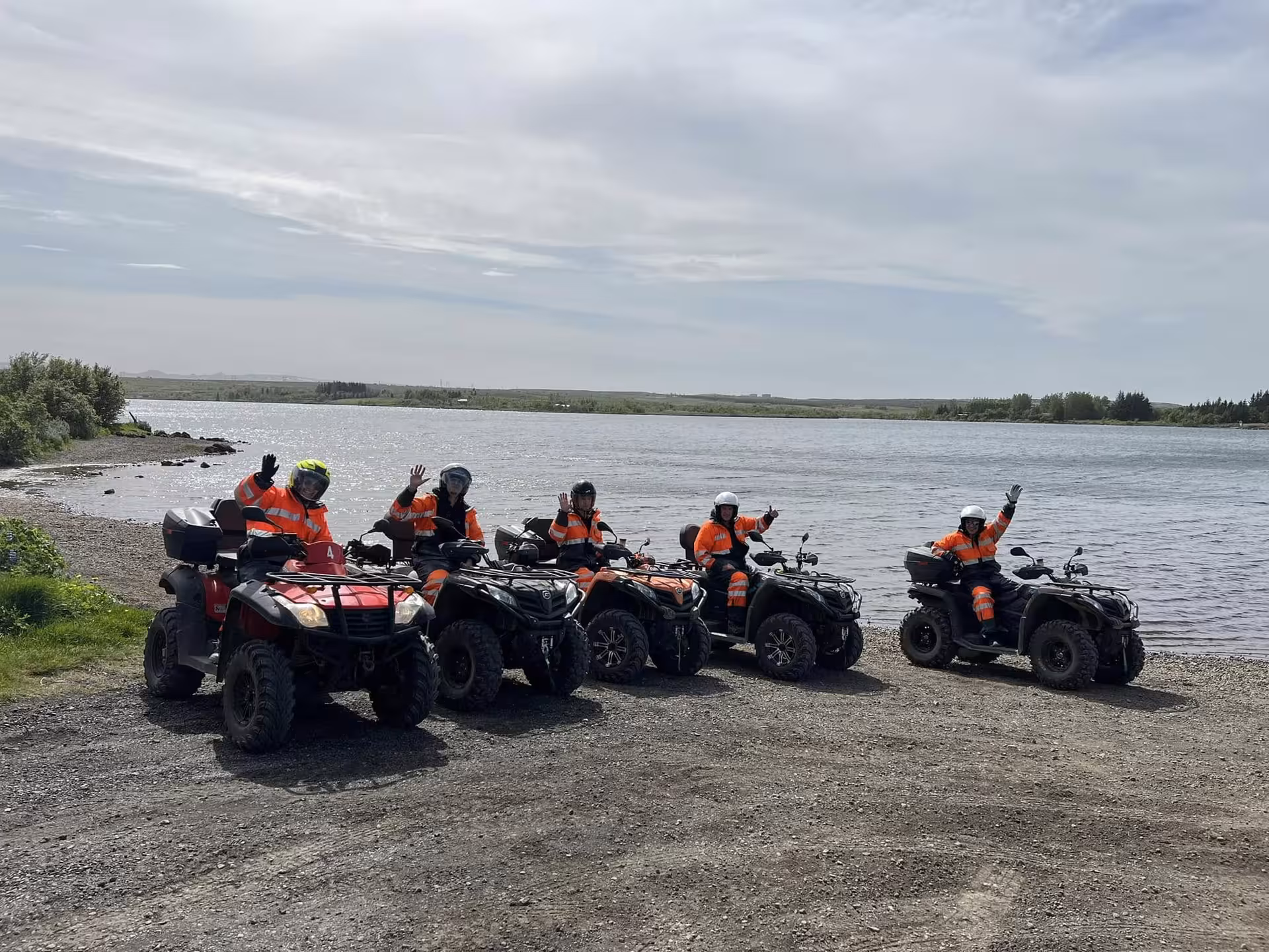 Group of five riders in orange safety gear on ATVs lined up along a lake shore in Iceland wilderness