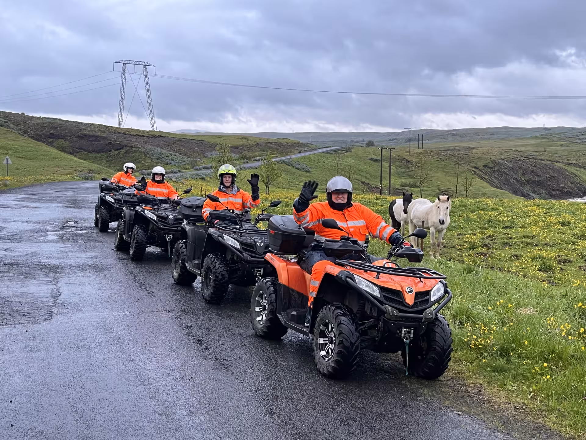 Four riders in orange safety gear on ATV quad bikes lined up on a wet road in Iceland's green wilderness with horses nearby