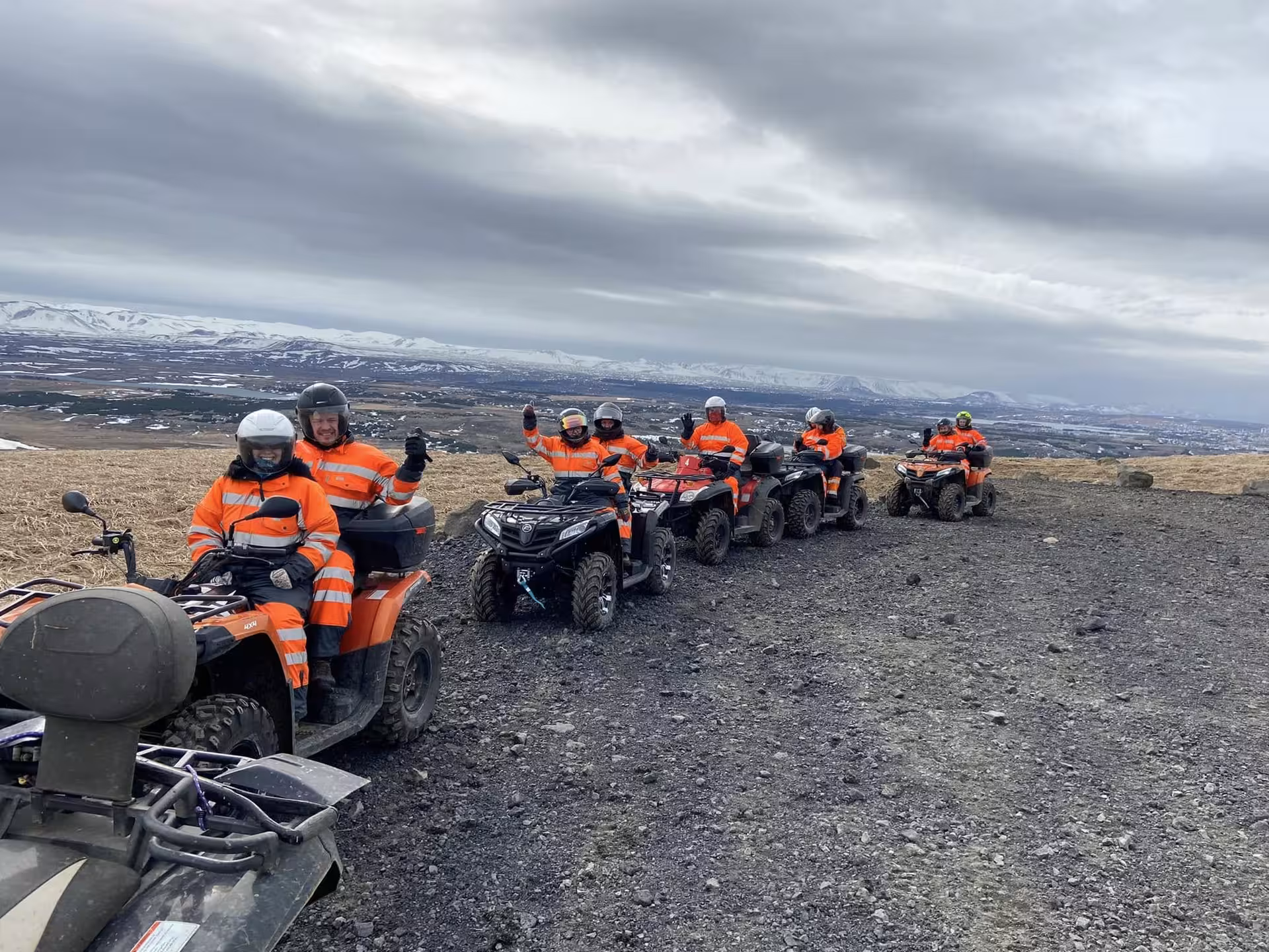 Group of tourists in orange gear riding ATV quad bikes on volcanic terrain near Reykjavik with snowy mountains behind