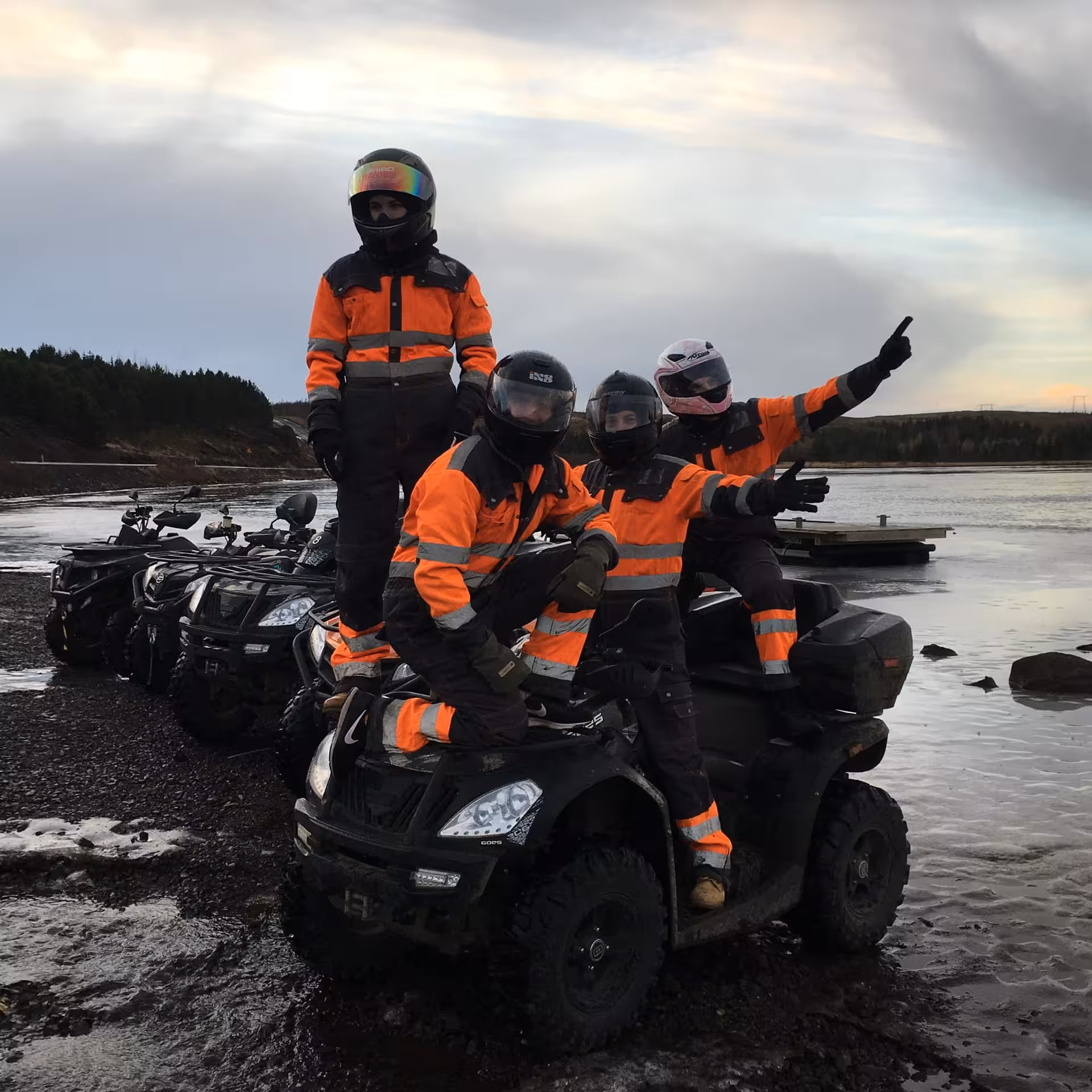 Four riders in orange safety gear posing on ATVs near a lake in Iceland wilderness at dusk