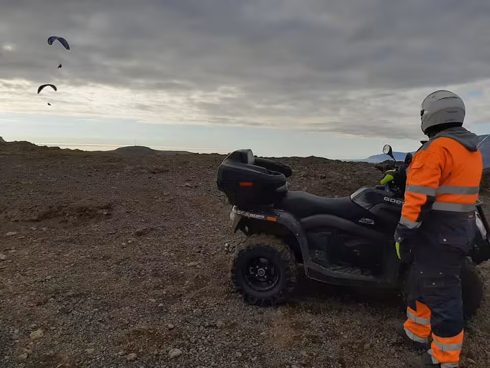 Rider in orange hi-vis gear beside ATV quad bike watching paragliders over Reykjavik wilderness at sunset