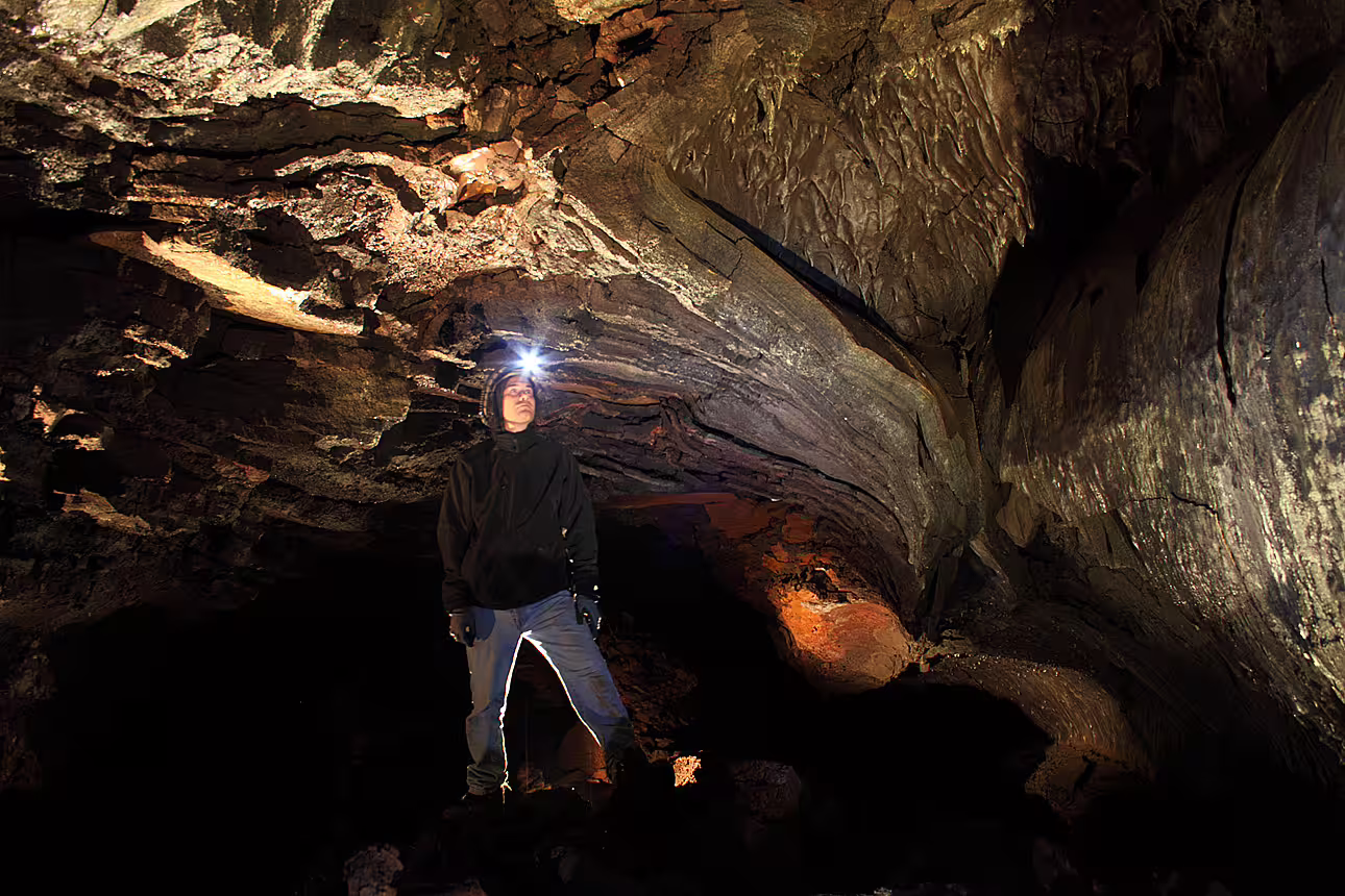 Explorer with headlamp standing inside dramatic lava cave tunnel with illuminated rock walls