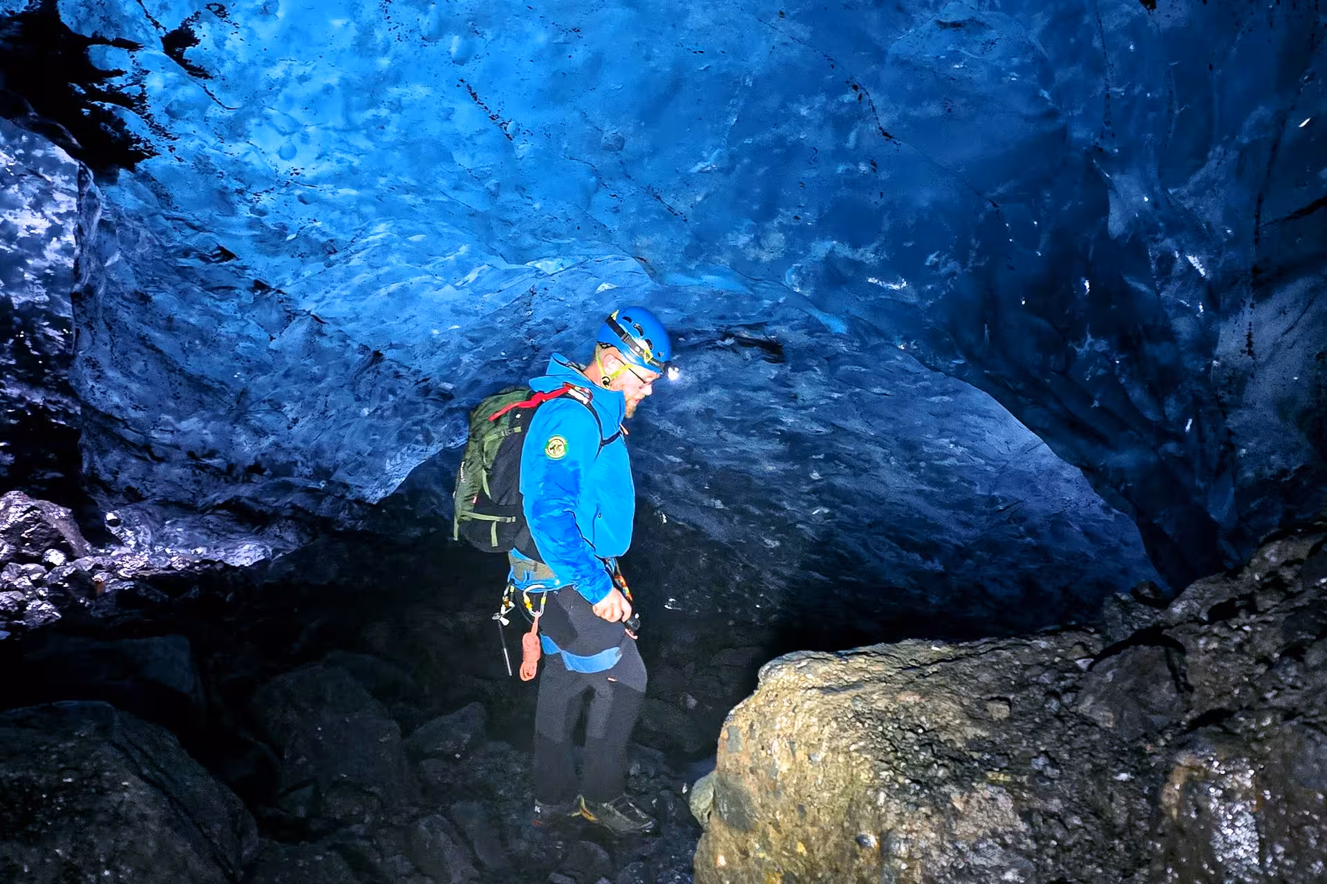 Explorer with blue helmet and headlamp navigating glowing blue ice cave tunnel in Iceland