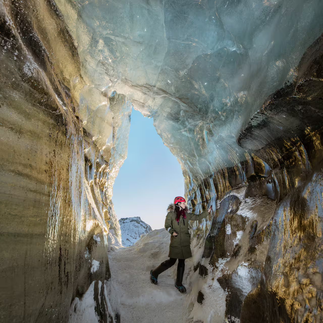 Explorer in red helmet stands inside Katla ice cave entrance with glacier walls and snowy Iceland mountains beyond