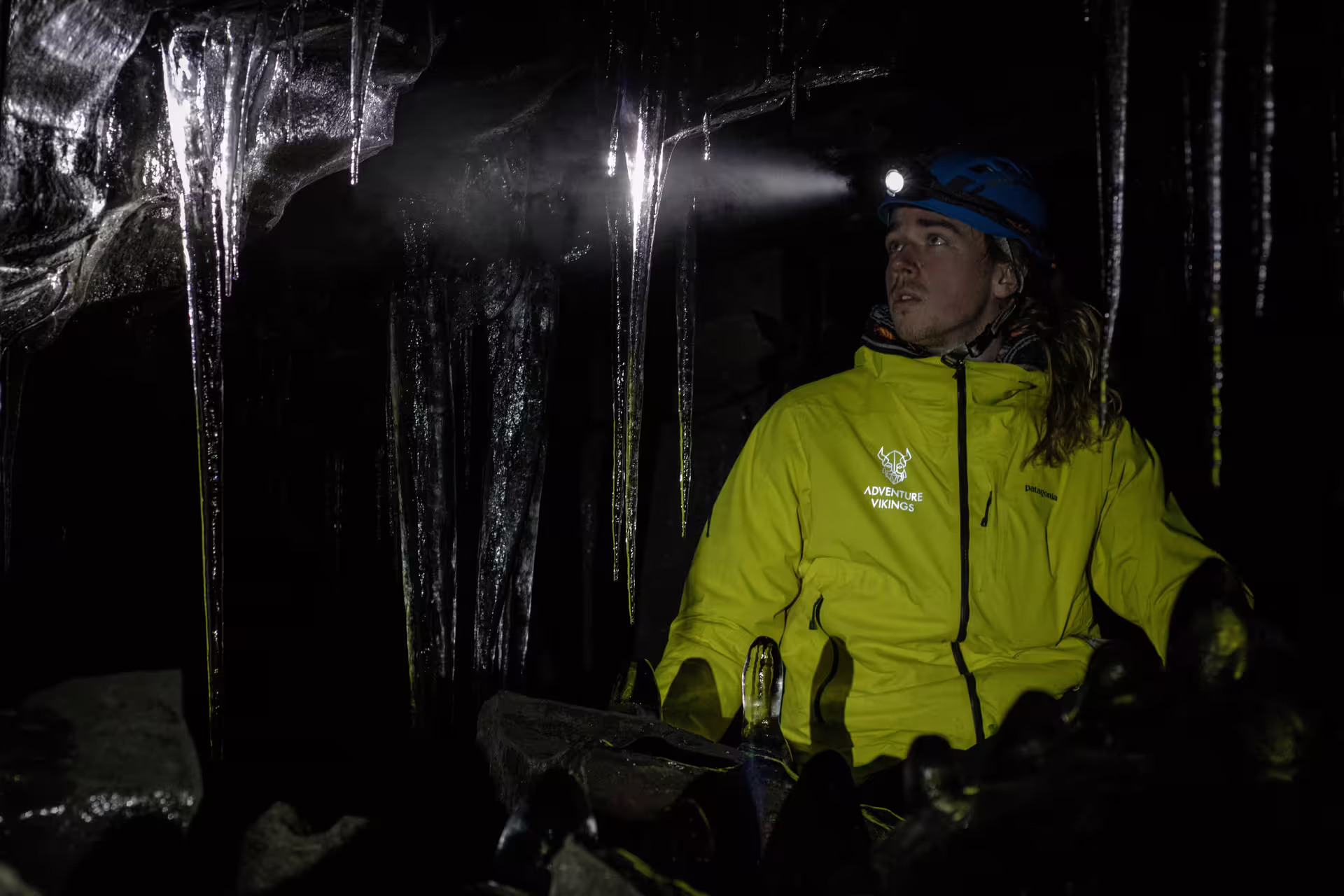 Explorer in yellow jacket and helmet with headlamp navigates icy formations inside Leidarendi Lava Tunnel on a small group tour.