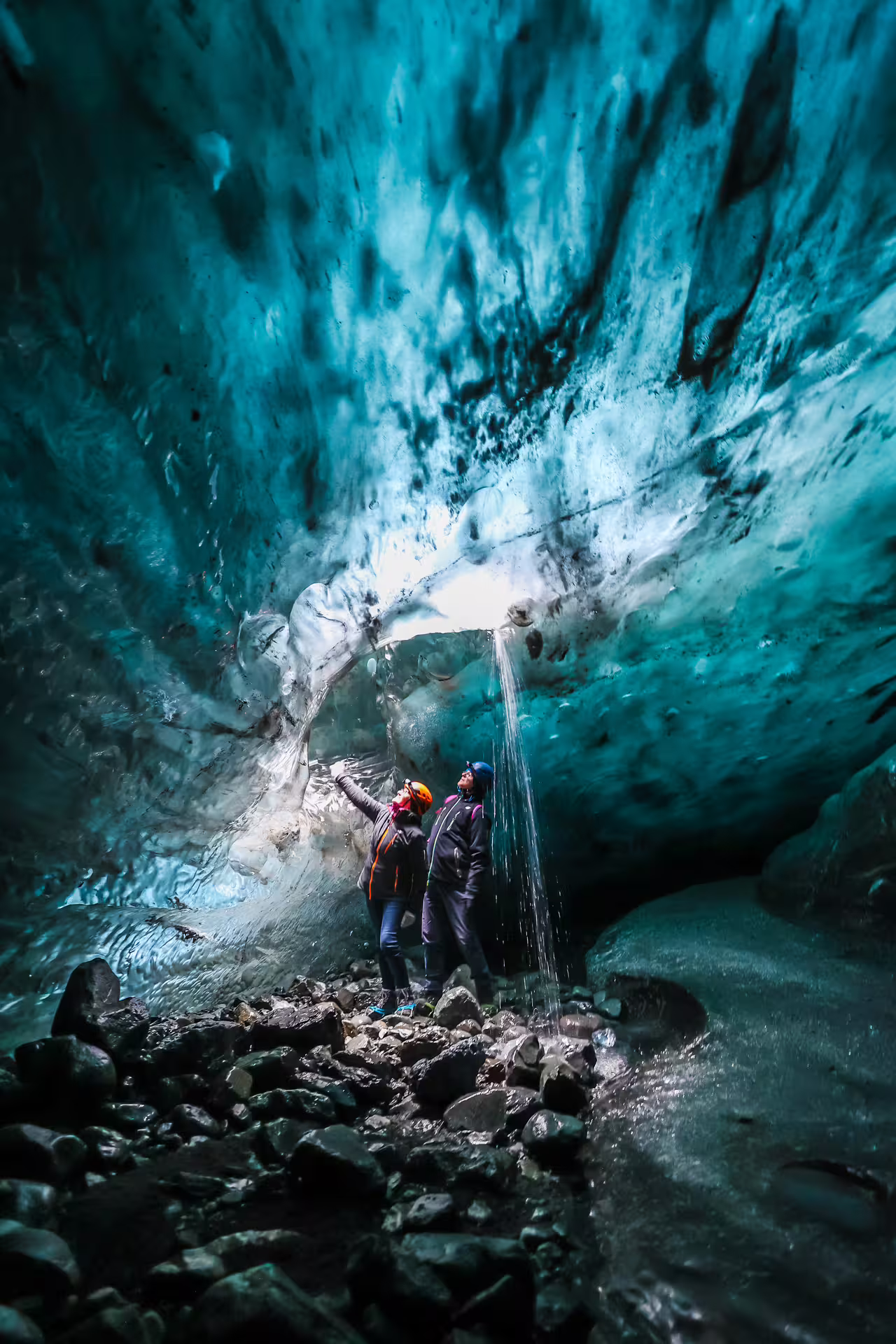 Two explorers wearing helmets marvel at a cascading waterfall inside a vivid blue Iceland glacier ice cave