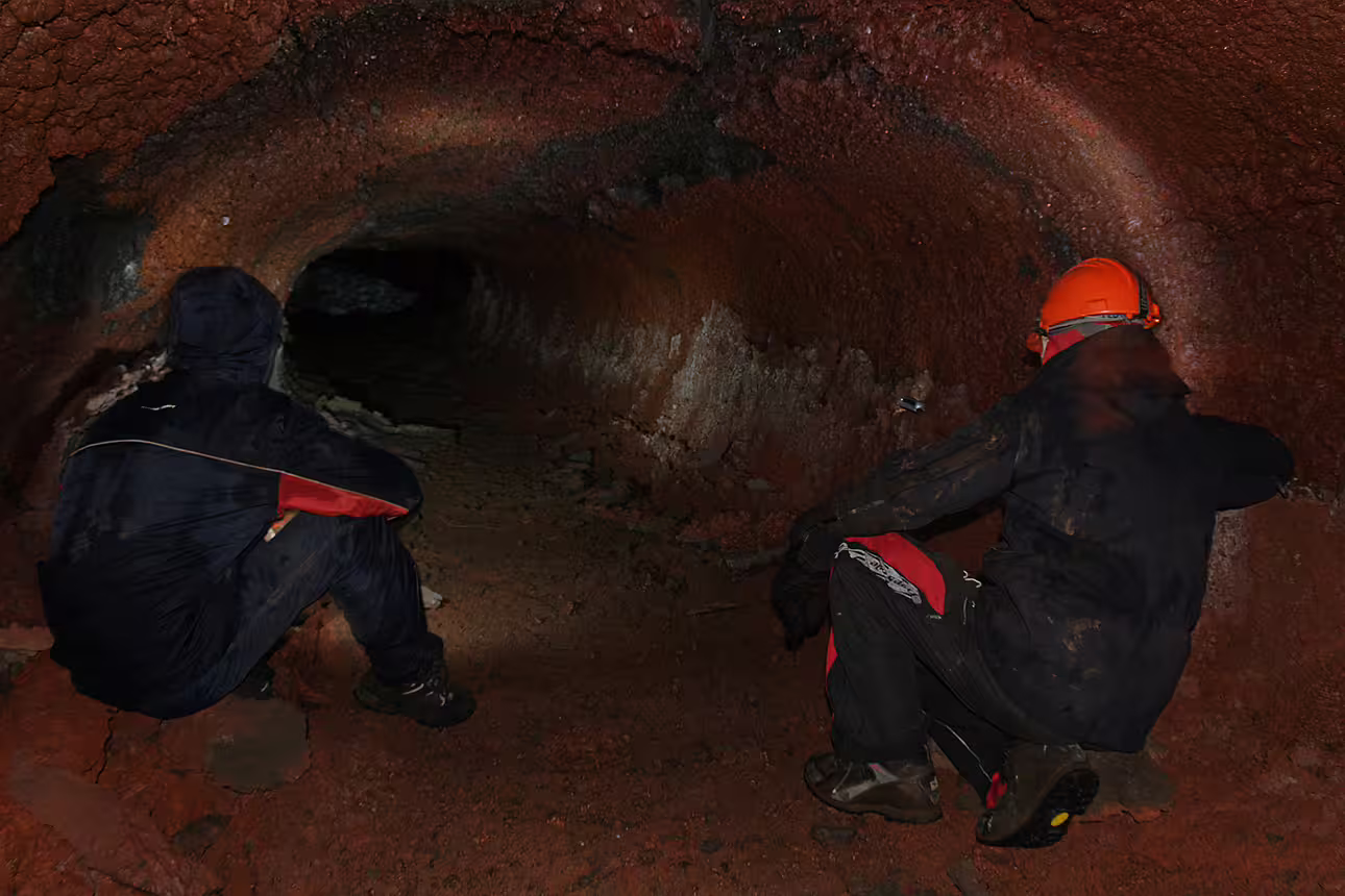 Two explorers crouching inside a red volcanic lava cave tunnel during a guided underground tour