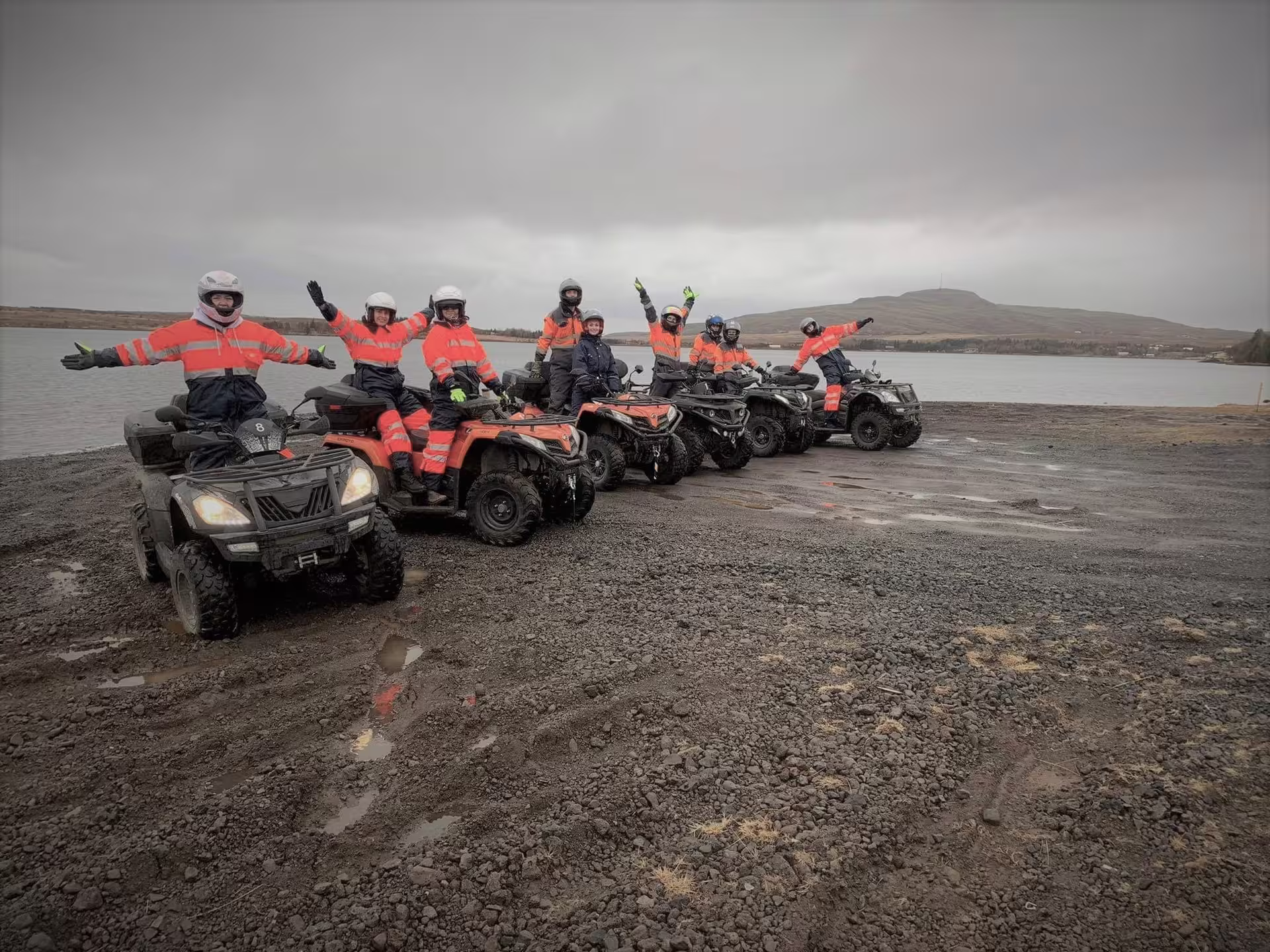 Group of riders in orange safety gear posing on ATVs near a lake in Reykjavik on a cloudy day