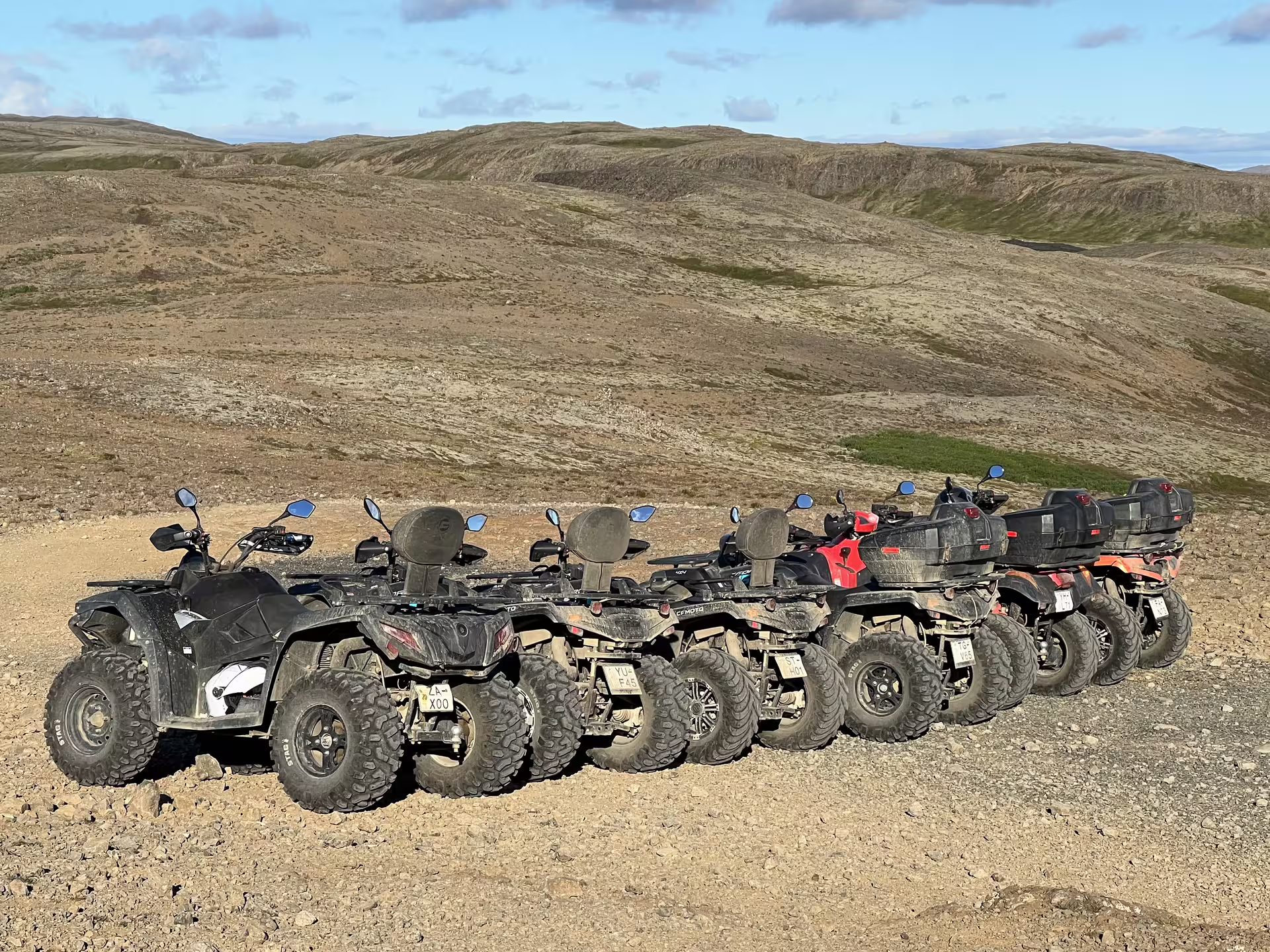 Row of muddy ATVs parked on gravel terrain with scenic Icelandic highland hills in background