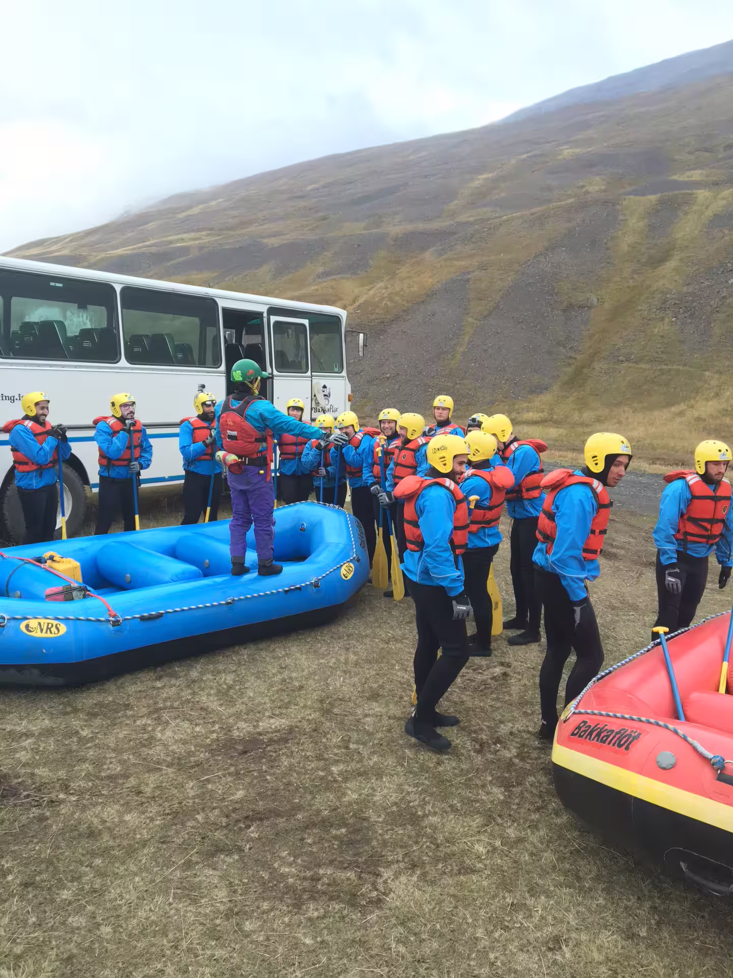 Group of adventurers in gear preparing for extreme river rafting beside colorful rafts and a bus on a scenic hillside.