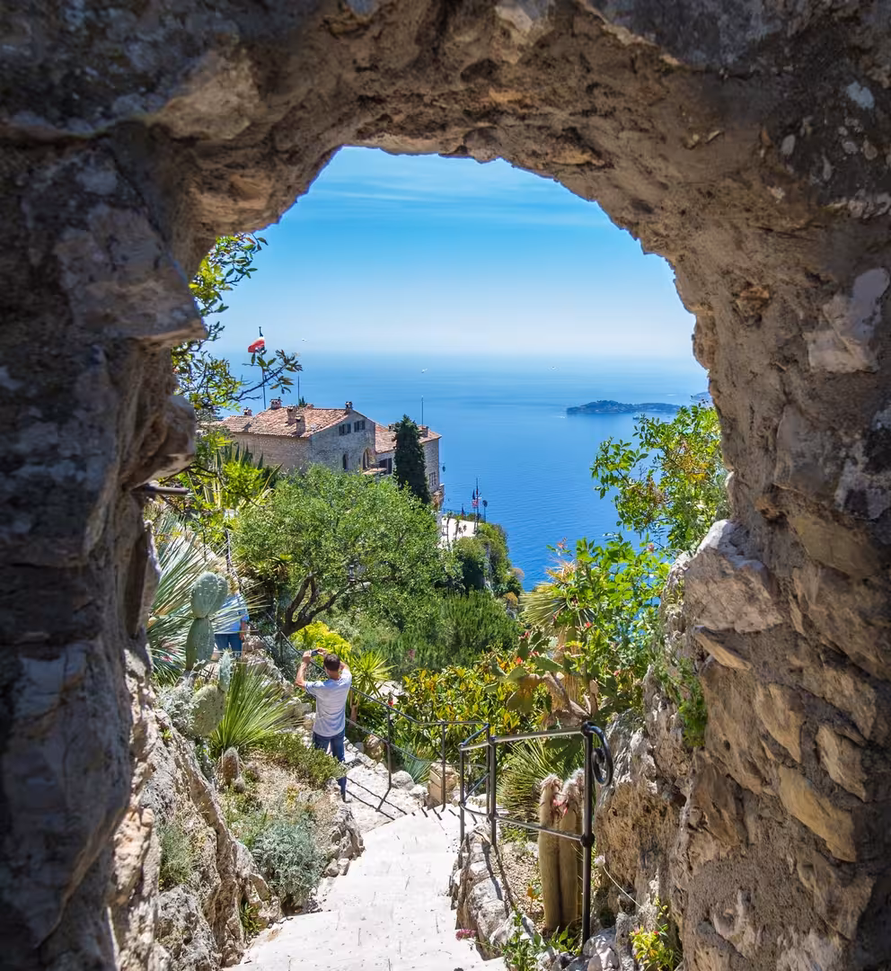 Stone arch gateway framing steps down to Eze Village garden with Mediterranean Sea and Monaco coastline beyond