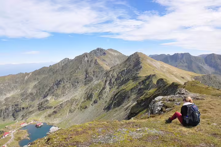 Hiker resting on Fagaras mountain ridge overlooking glacial lake and dramatic rocky peaks, Transfagarasan Romania