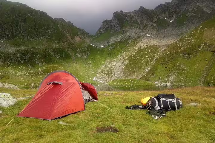 Red camping tent pitched on green alpine meadow with mountain lake and rocky Fagaras peaks in background