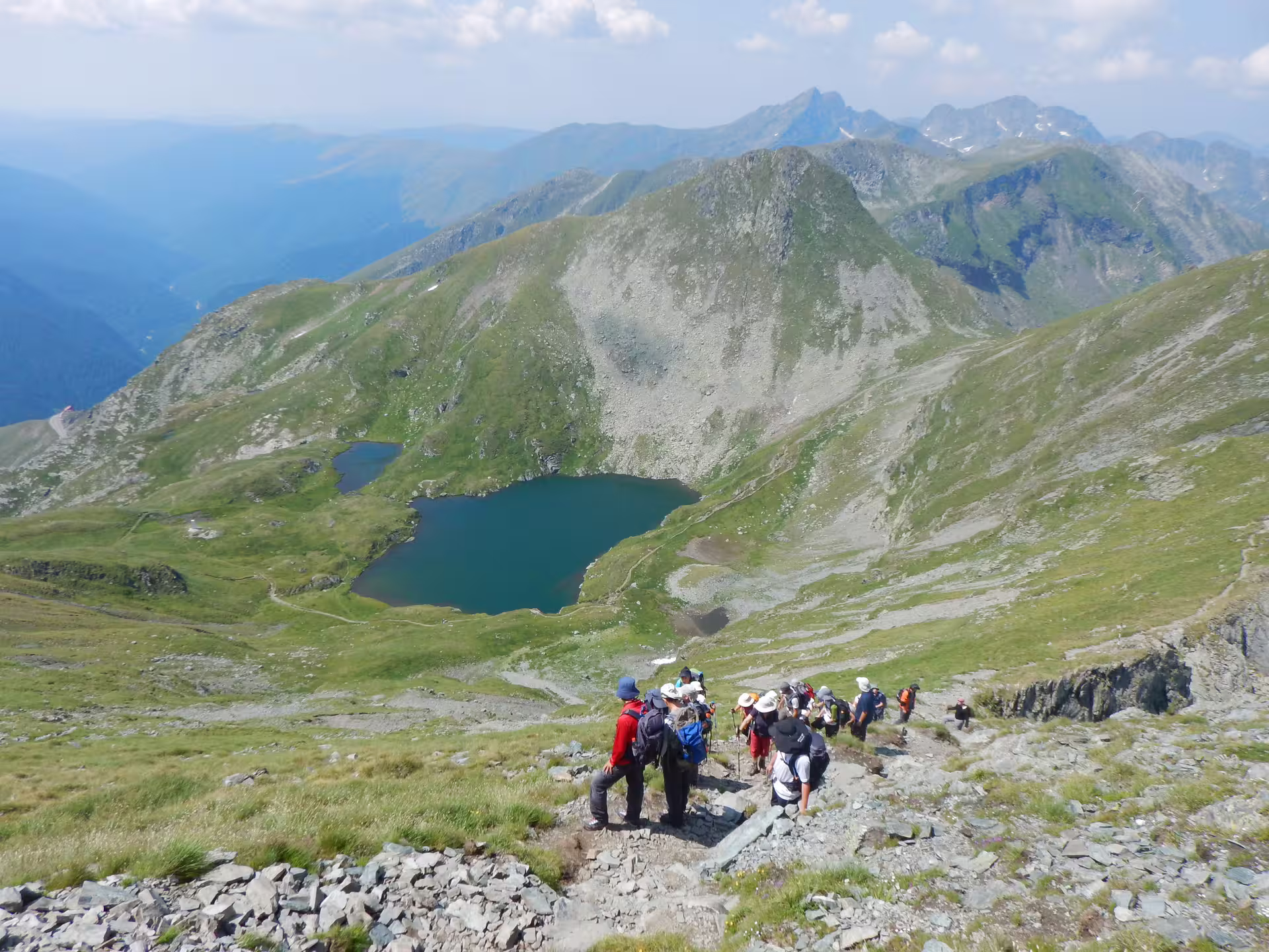 Hikers trekking down rocky trail in Fagaras Mountains with alpine lake and peaks in background