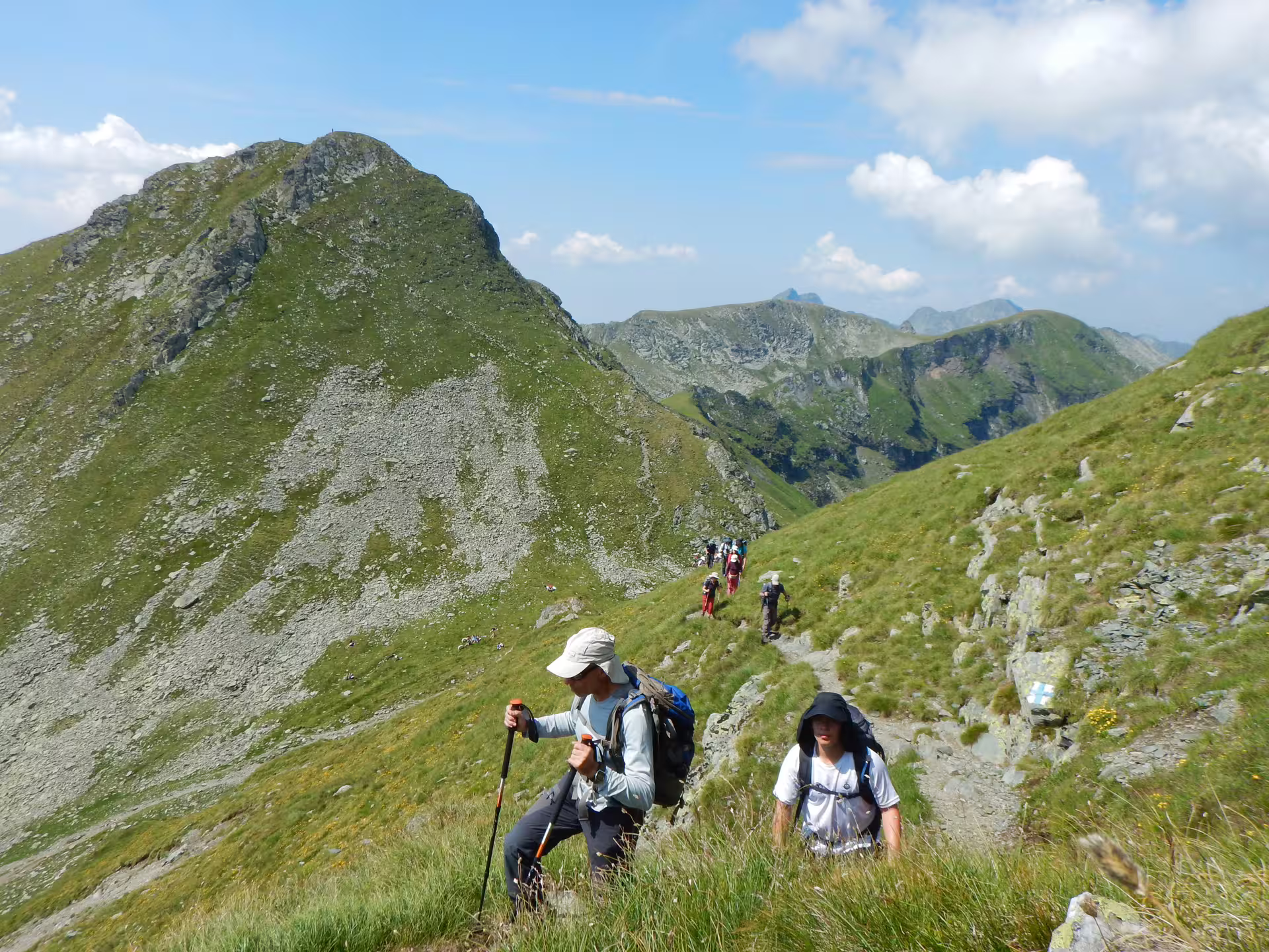 Hikers trekking up a mountain ridge trail in the Fagaras Mountains with peaks in background