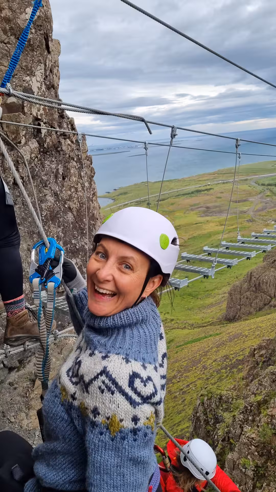 Smiling climber in Nordic sweater and white helmet on Falcon Rock Via Ferrata cliff face near Reykjavik