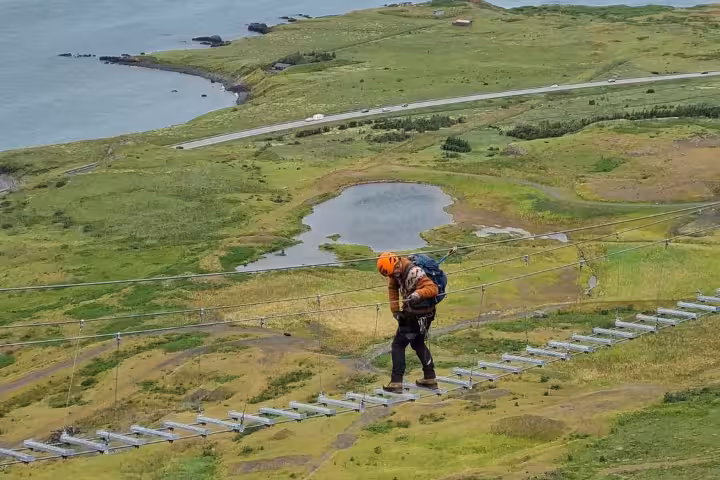 Climber crossing suspended rope bridge on Via Ferrata route above green Icelandic landscape near Reykjavik
