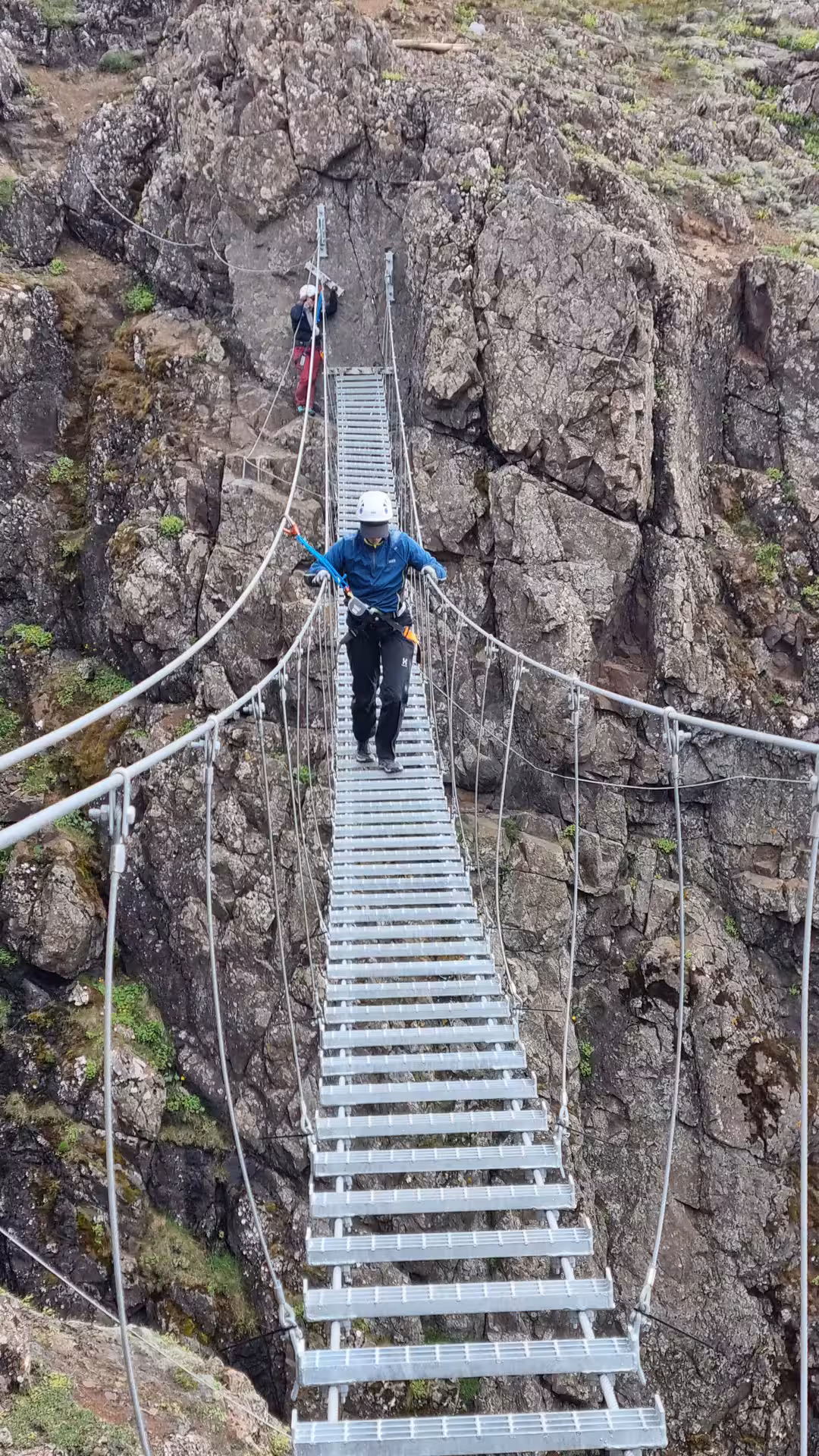 Climber in blue jacket and white helmet crossing metal suspension bridge on via ferrata route in Iceland
