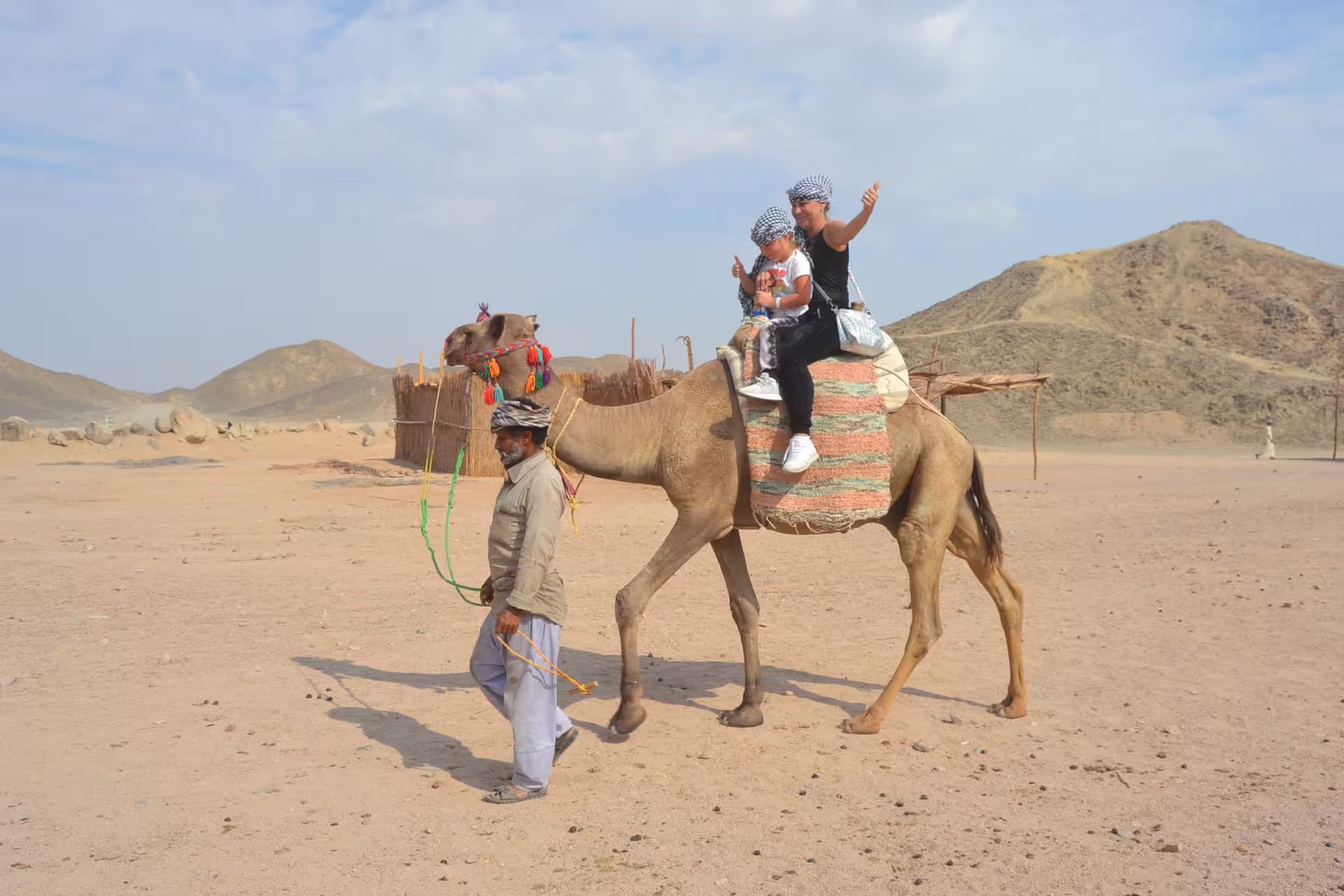 Family camel ride at Bedouin village stop on Hurghada quad safari, Egypt, with desert hills behind