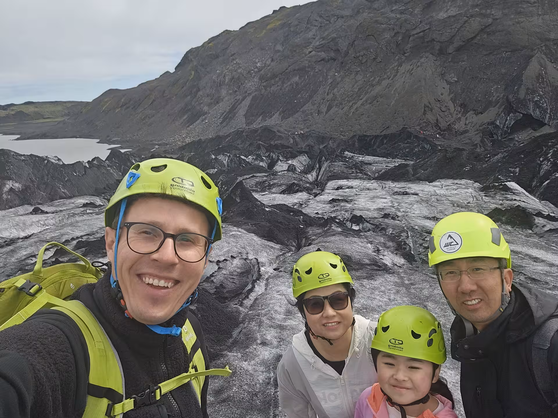 Tour guide and family wearing green helmets posing on a dark glacier with volcanic landscape and lake in background
