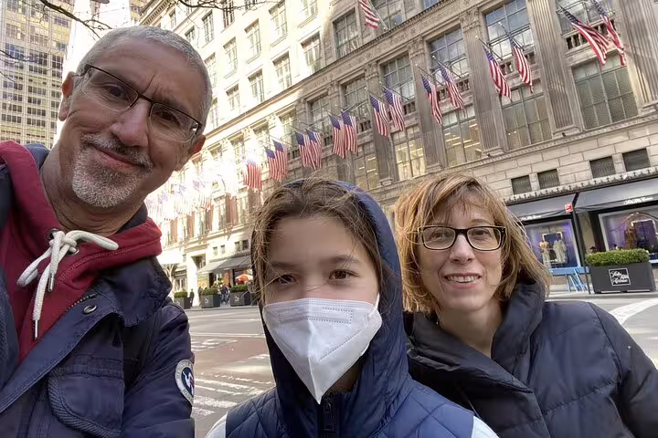 Family of three posing for selfie on Midtown Manhattan street with American flags and historic building behind
