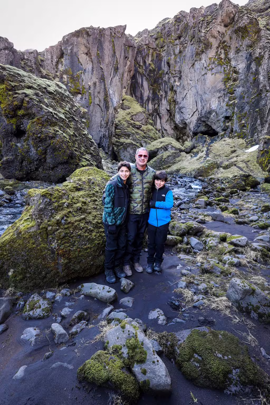 A family of three poses in a rugged moss-covered Icelandic canyon with black sand and towering rock walls on a grand Iceland tour.