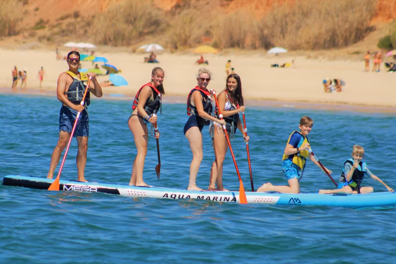 Smiling family group enjoying a Mega Paddle rental on a giant SUP board in calm blue sea near a sunny sandy beach