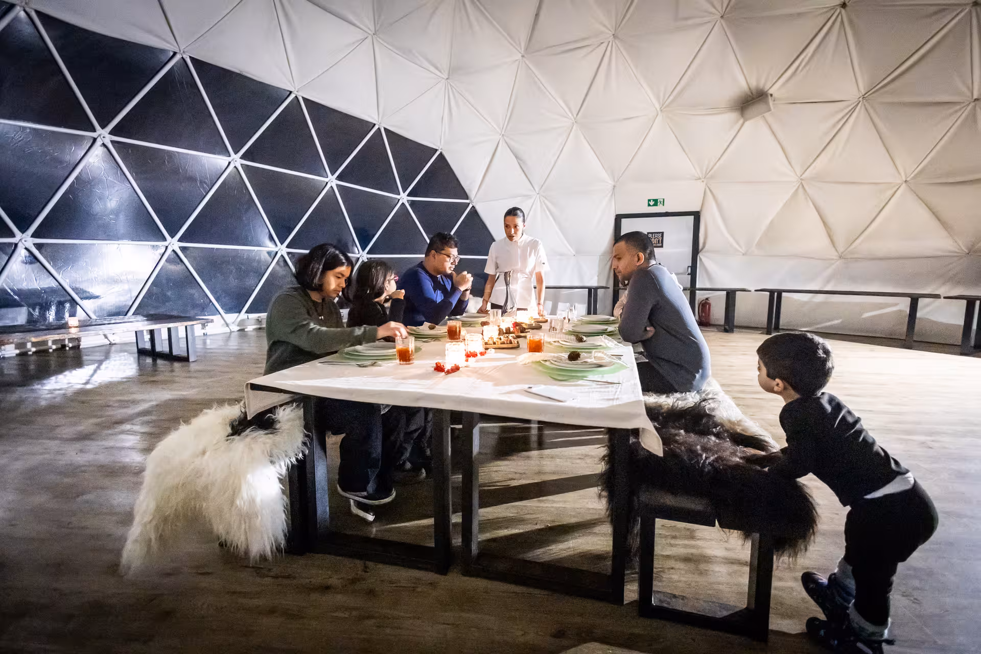 Family enjoying a cozy northern lights dinner inside a geodesic dome with fur-draped benches in Iceland
